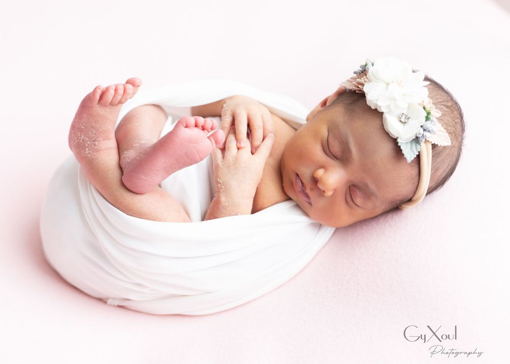 Newborn photography session featuring baby wrapped in soft white fabric against pink backdrop.