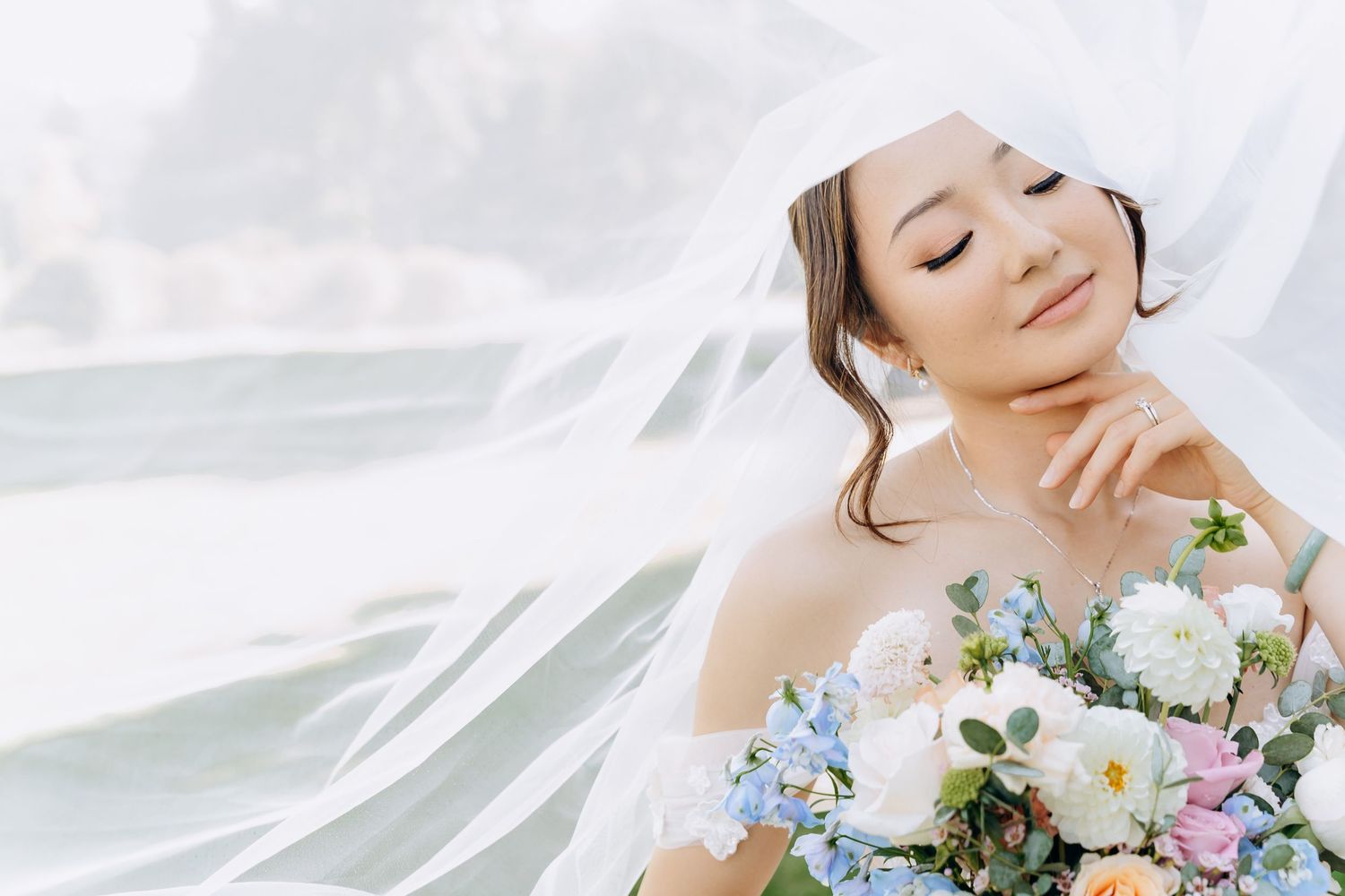 Bride in off-shoulder dress holds pastel bouquet with white and blue flowers as veil flows in breeze.