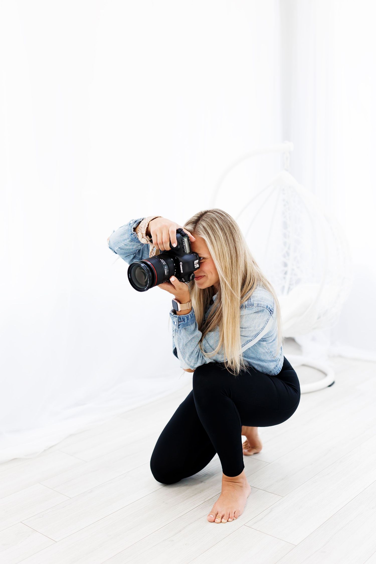 Photographer in denim shirt crouches low with professional camera in bright white studio setting.