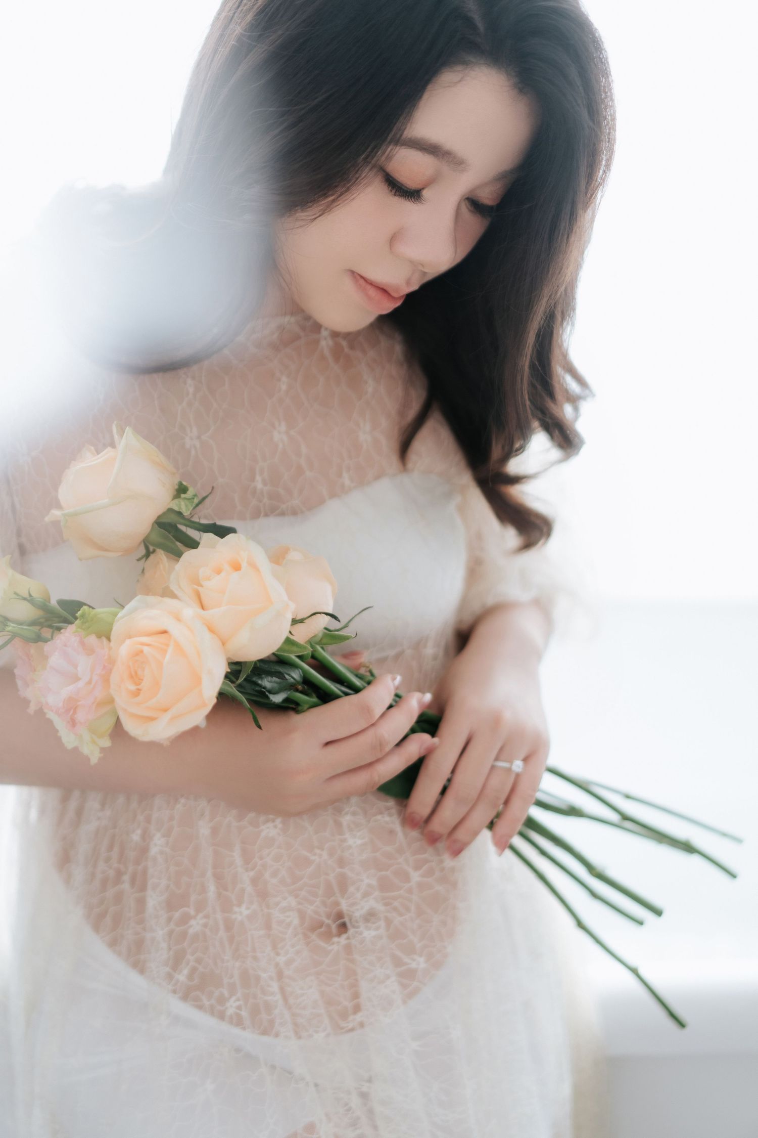 Young Asian woman in white lace dress holding peach roses bouquet, looking down gracefully in soft natural light.一位年輕的亞洲女子身穿白色蕾絲連身裙，手捧一束桃色玫瑰