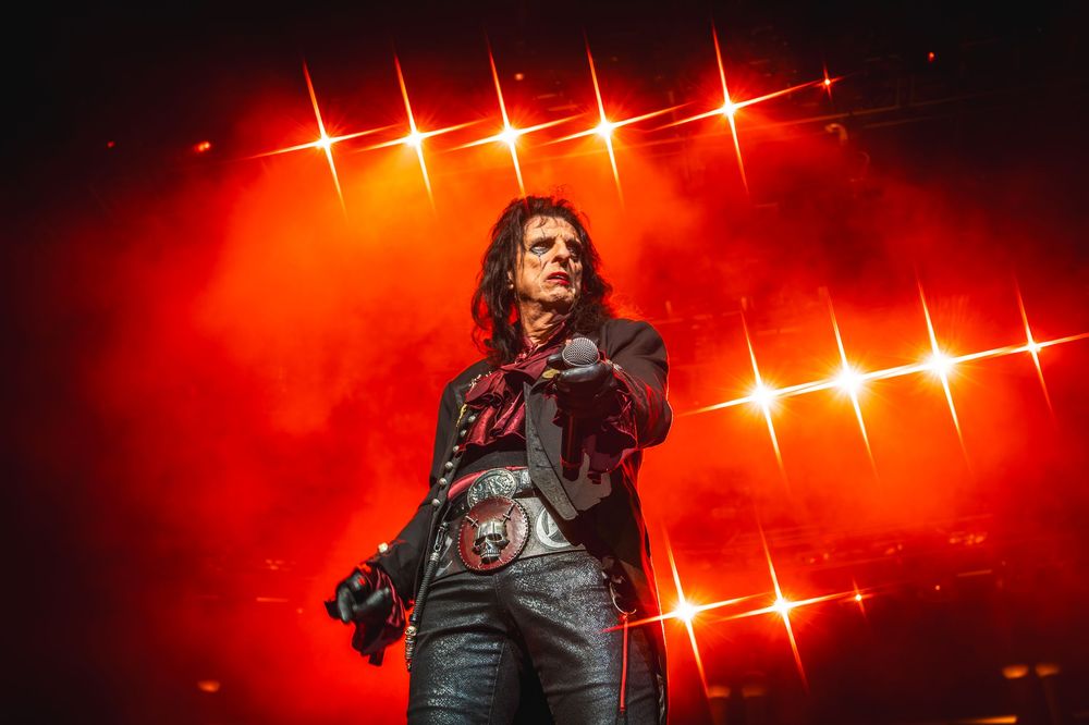 A performer in black clothing sings passionately on stage with dramatic red lighting and star-like effects in the background.