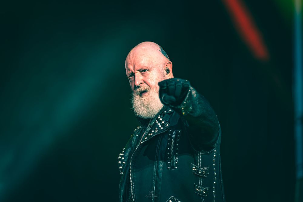 Concert performer in black clothing singing on stage with dramatic teal lighting and atmospheric effects in the background.