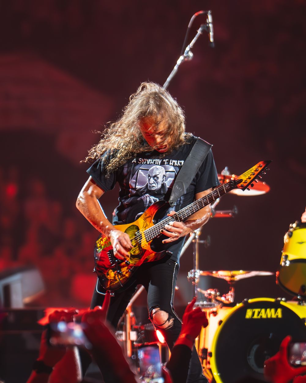 A long-haired guitarist performs energetically on stage with a colorful guitar in front of a red backdrop and Tama drum kit.