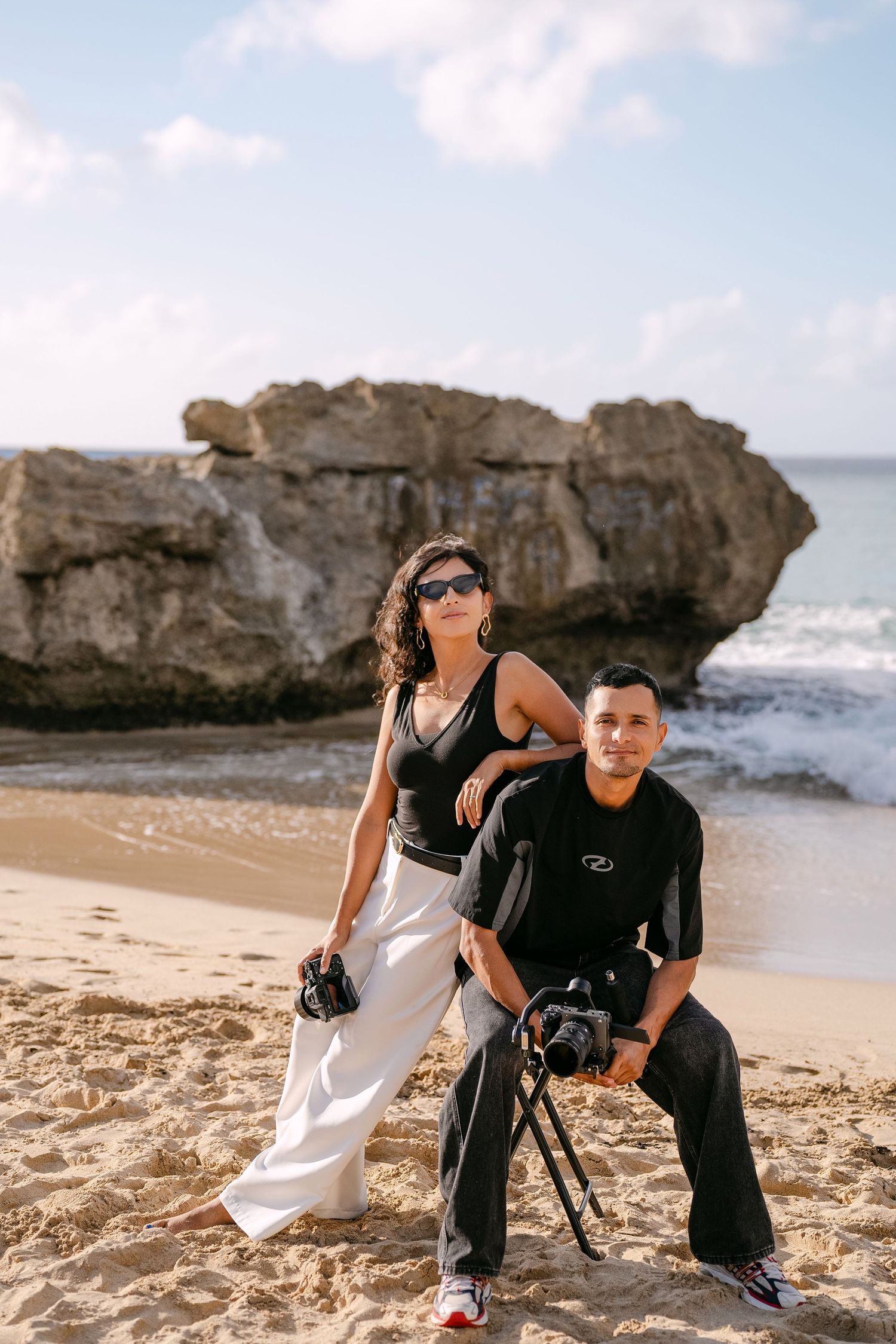 Two photographers with camera equipment pose together on a scenic beach with rocky cliffs in the background.