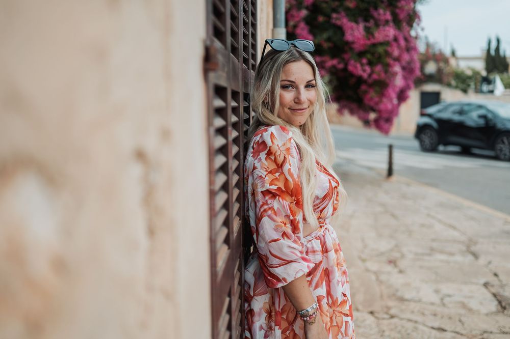 A person in a floral orange and white dress poses against a wall with pink bougainvillea flowers in the background.