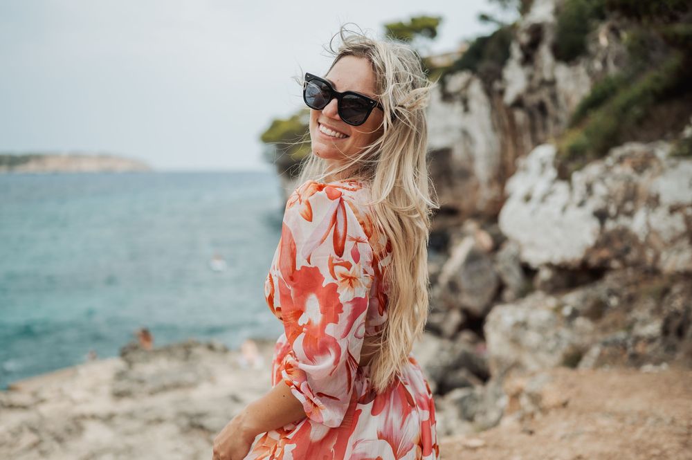 Woman in coral floral dress and sunglasses poses on rocky Mediterranean coastline with turquoise water view.