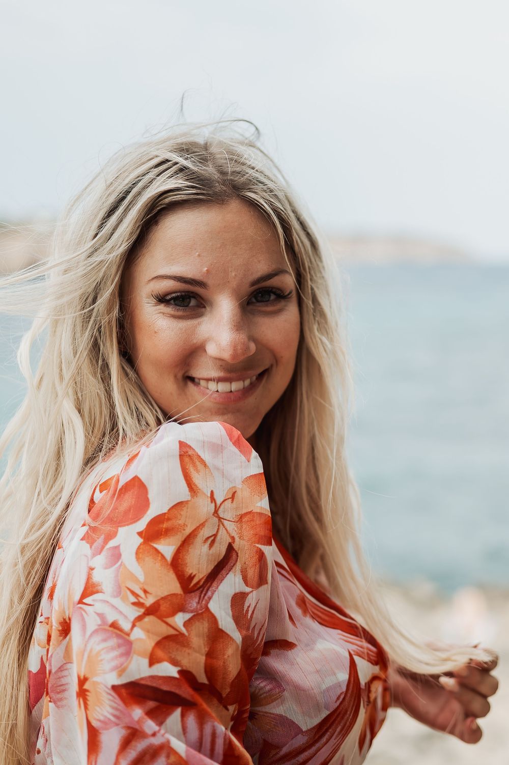 A person in a coral floral dress poses against a coastal background with blue water and rocky terrain.