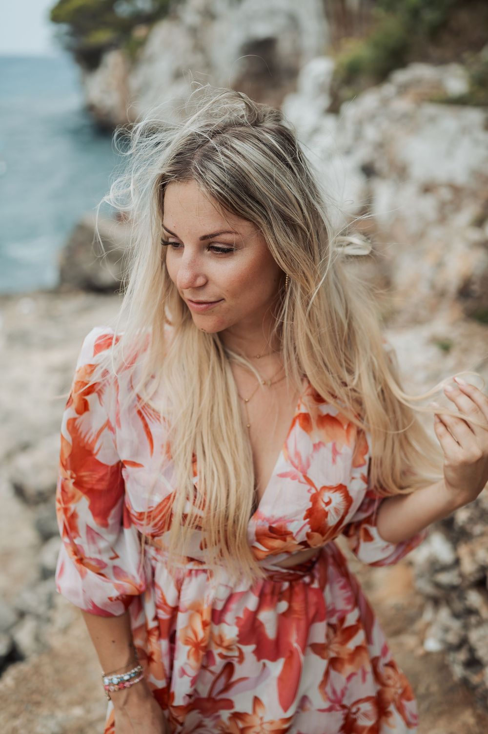 A woman in a floral coral dress stands near rocky coastal cliffs with windswept blonde hair on a sunny day.
