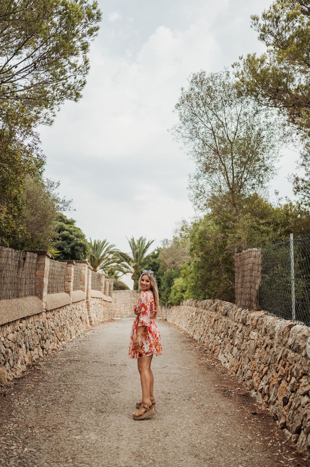 Woman in floral dress stands on stone-walled path lined with palm trees and Mediterranean foliage.