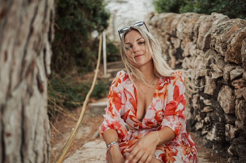 Woman in floral dress poses outdoors against rustic stone wall in natural lighting.