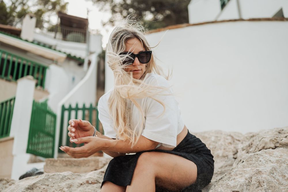 Woman in casual attire sits on rocky ledge against white Mediterranean building wall.
