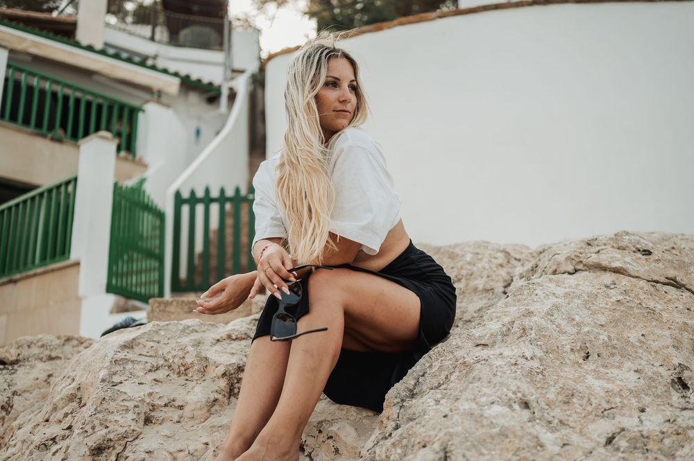 A person in a white top and dark shorts sits on rocky terrain near white Mediterranean-style buildings.