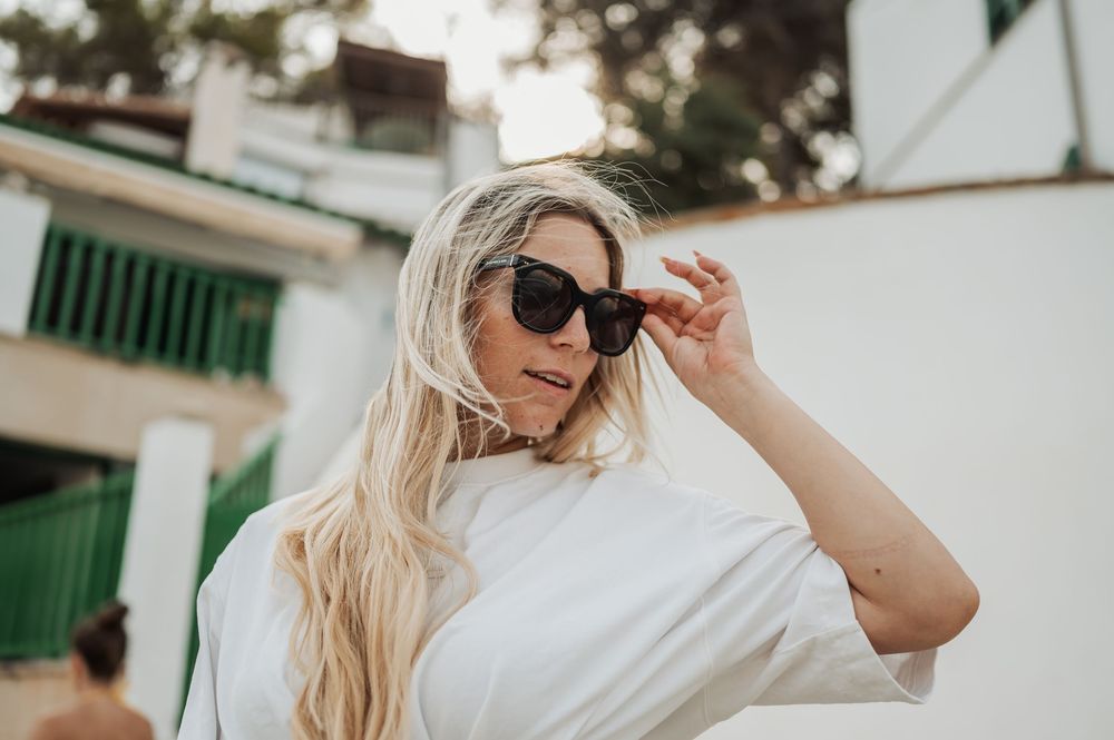A person in sunglasses and white clothing adjusts their frames while posing outdoors against a building with green accents.