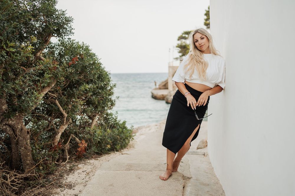 A woman in a white top and black skirt leans against a white wall along a coastal path with ocean views.