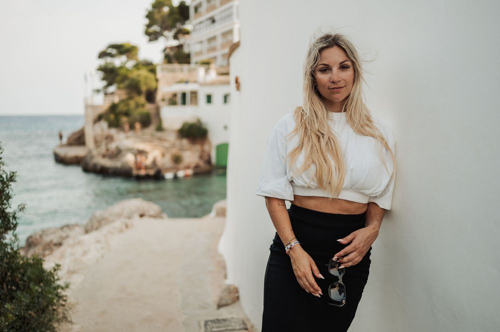 A person in a white crop top and black skirt poses against a wall near a Mediterranean coastal villa and beach.