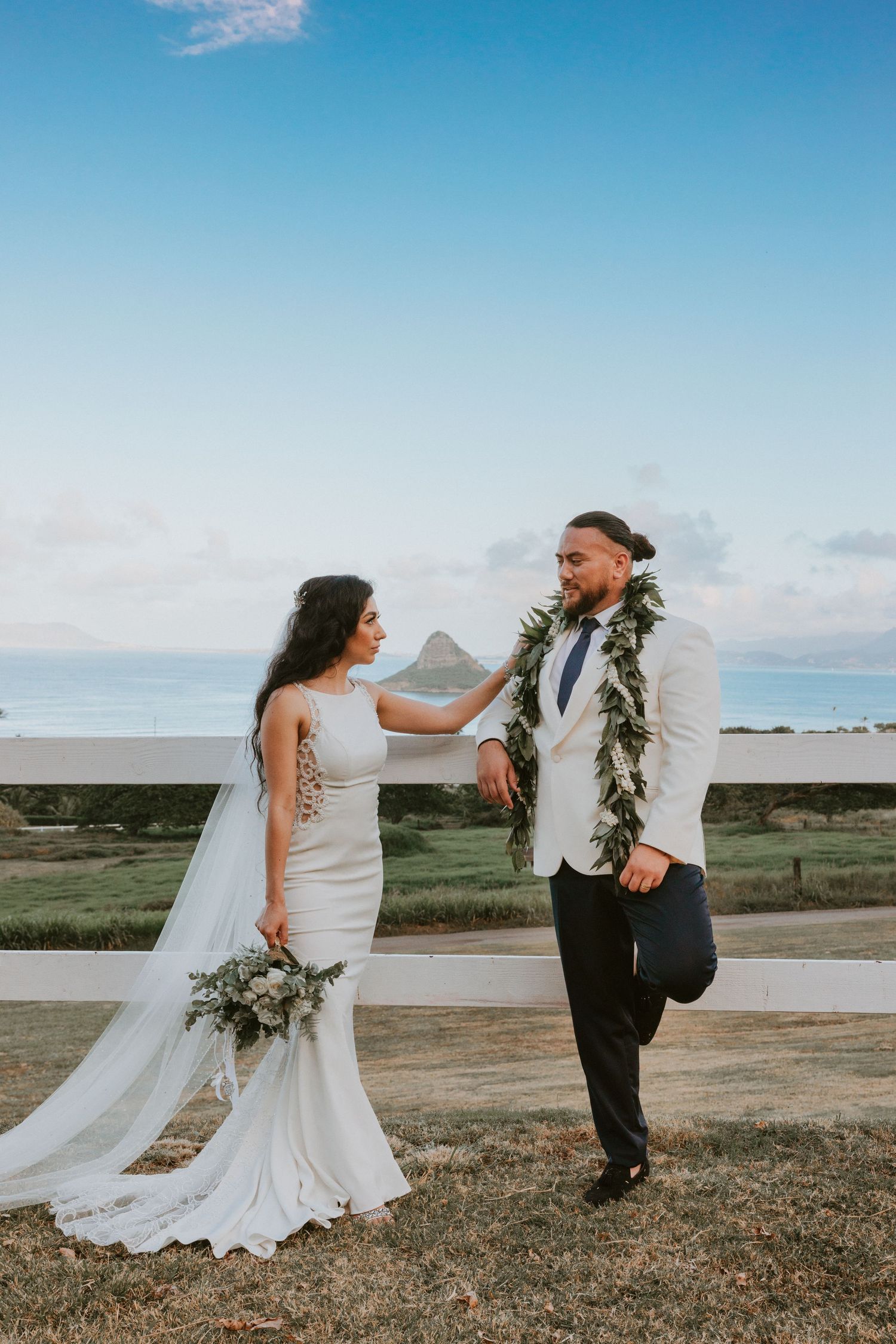Hawaiian wedding couple share a playful moment by white fence with ocean and mountain views.