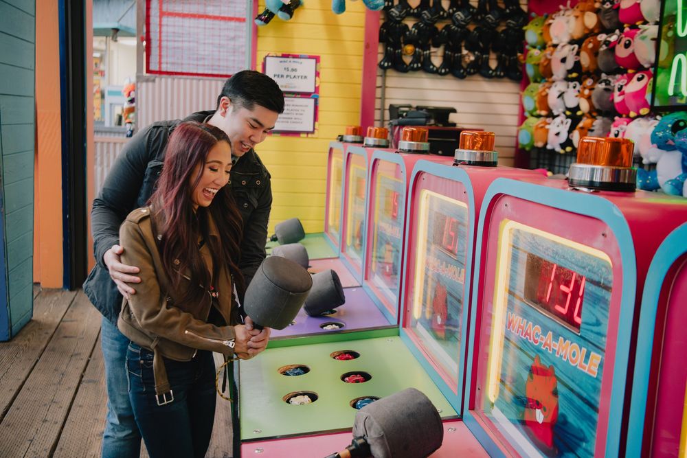 Couple playing whack-a-mole arcade game surrounded by colorful prize stuffed animals.