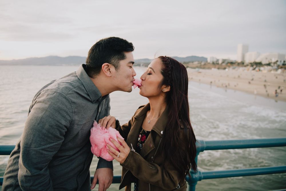 Romantic couple sharing a kiss over pink cotton candy on a beach pier at sunset.