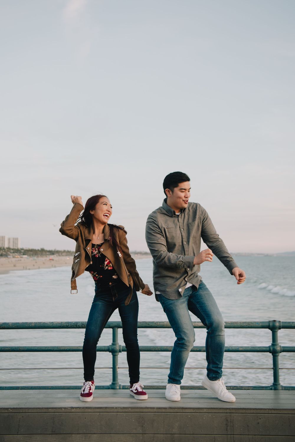 Joyful couple dancing together on a pier railing with ocean beach in background.