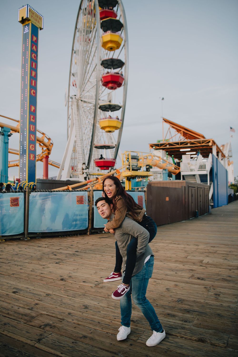 Happy couple doing a piggyback ride at Santa Monica Pier with Ferris wheel behind them.