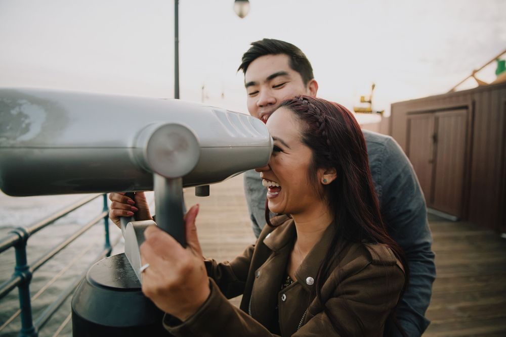 Laughing couple using a coin-operated telescope together on a seaside pier.
