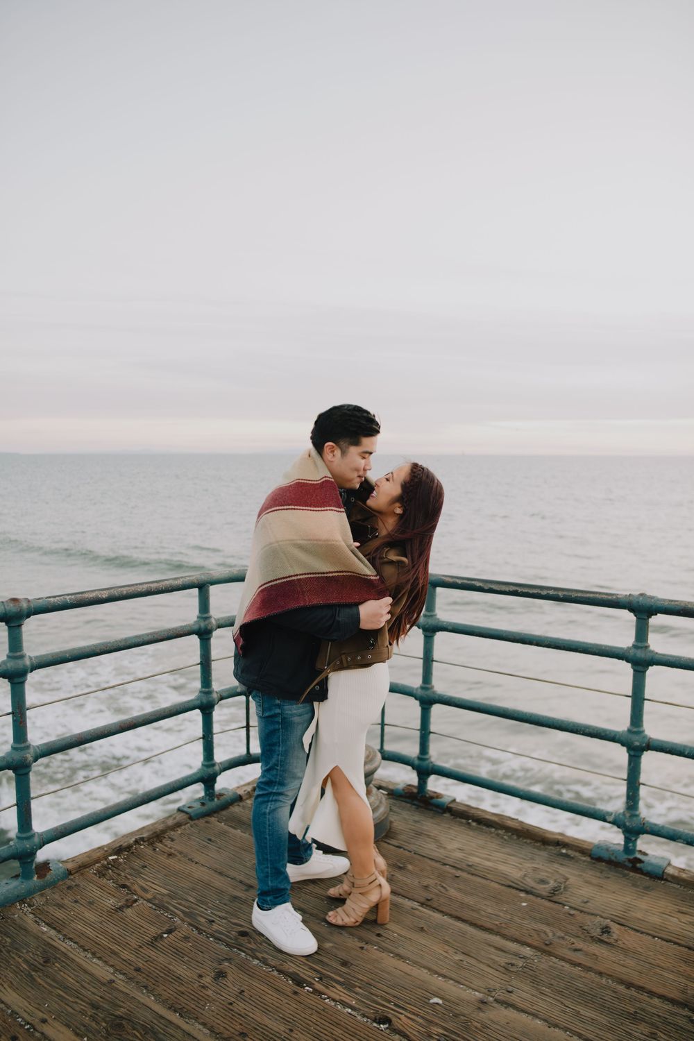 Couple wrapped in a cozy blanket sharing a tender kiss on an ocean pier at dusk.