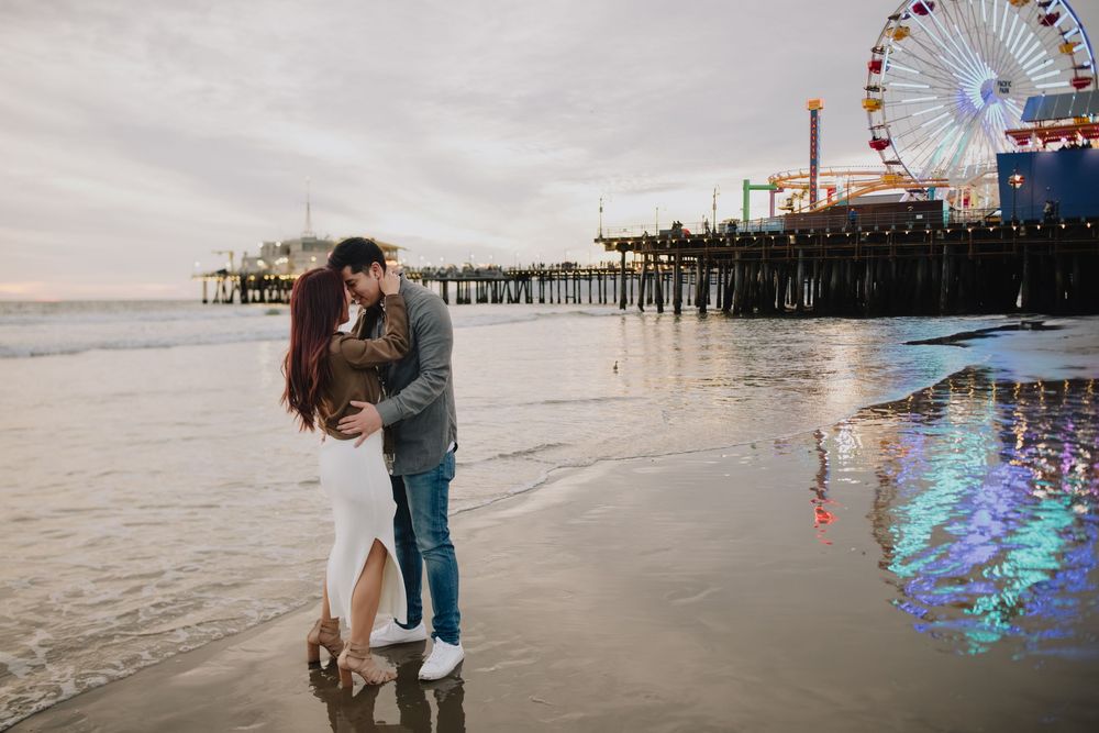 Couple kissing on Santa Monica beach at sunset with iconic pier and ferris wheel reflected in wet sand.