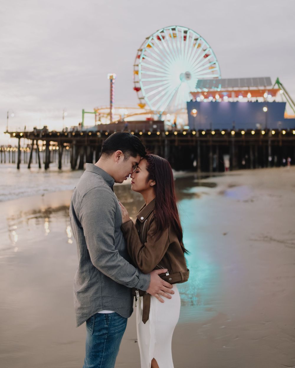 Romantic couple embracing on Santa Monica beach at dusk with illuminated ferris wheel glowing behind them.