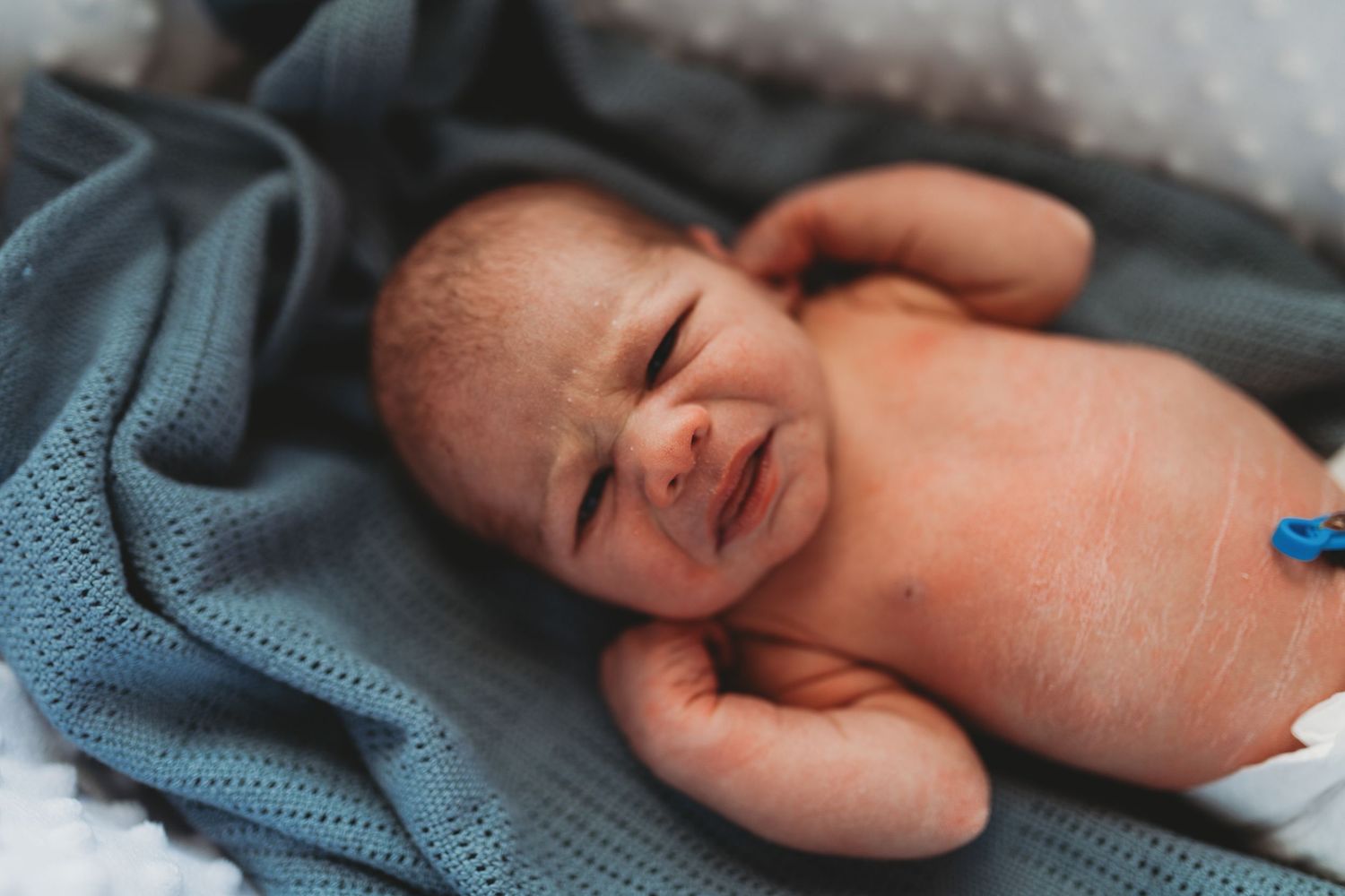 A newborn baby lies on soft blue blankets while crying and showing emotion.