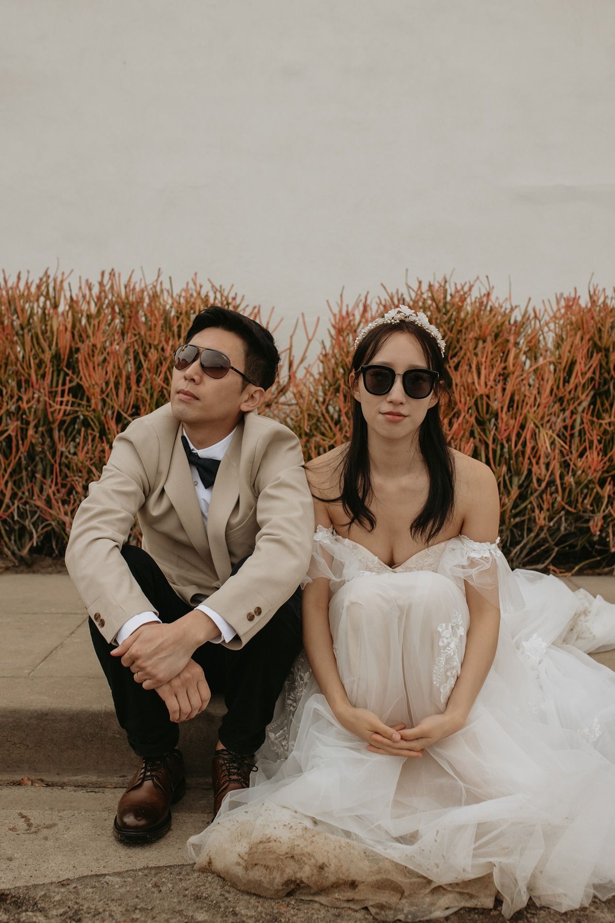 Stylish couple in sunglasses and wedding attire pose casually in front of tall orange plants during a photo session at Windansea Beach.