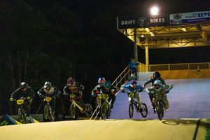 BMX riders compete at night on an illuminated dirt track with jumps under bright stadium lights.