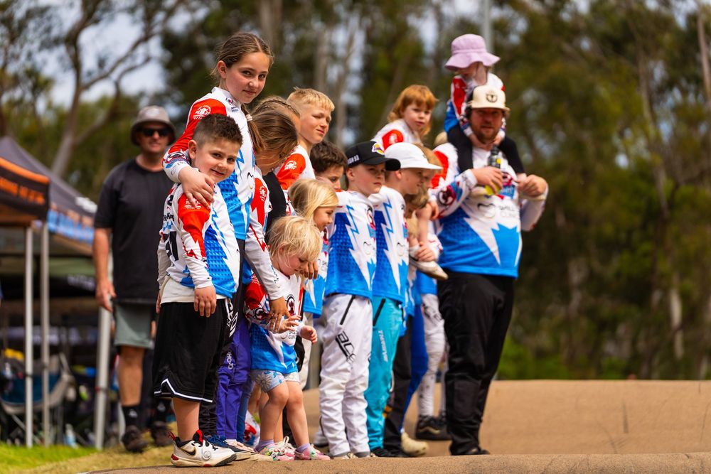 Athletes and spectators gather on a dirt field during a sporting event with trees in the background.