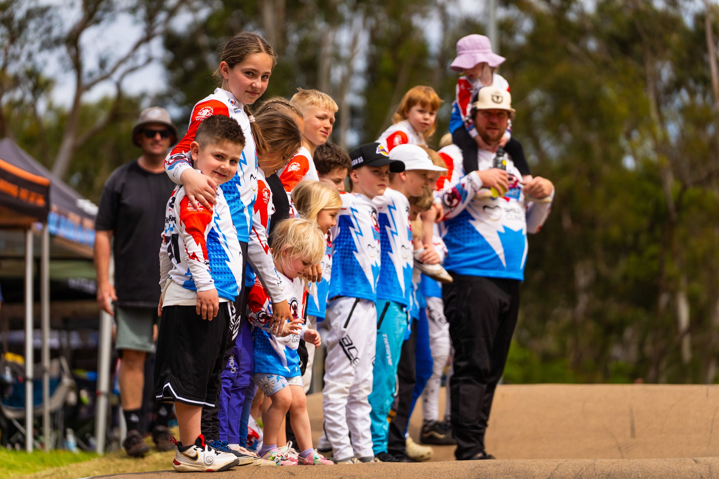 Athletes and spectators gather on a dirt field during a sporting event with trees in the background.
