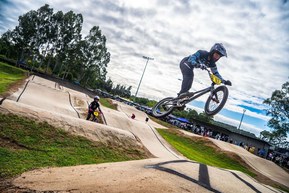 BMX rider performs aerial maneuver on dirt jump track with dramatic cloudy sky and trees in background.