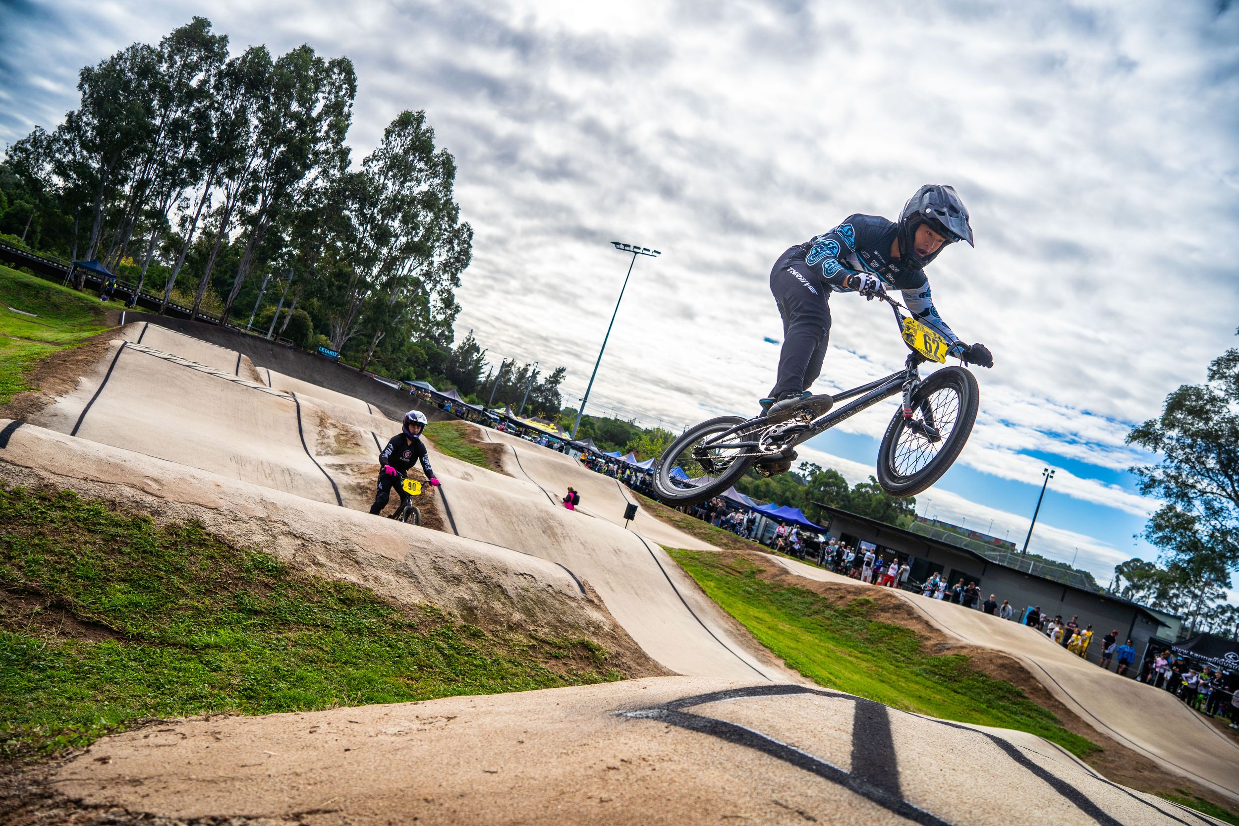BMX rider performs aerial maneuver on dirt jump track with dramatic cloudy sky and trees in background.