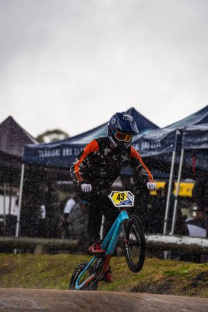 Mountain biker performs stylish aerial maneuver on dirt jump track during competition in overcast weather.