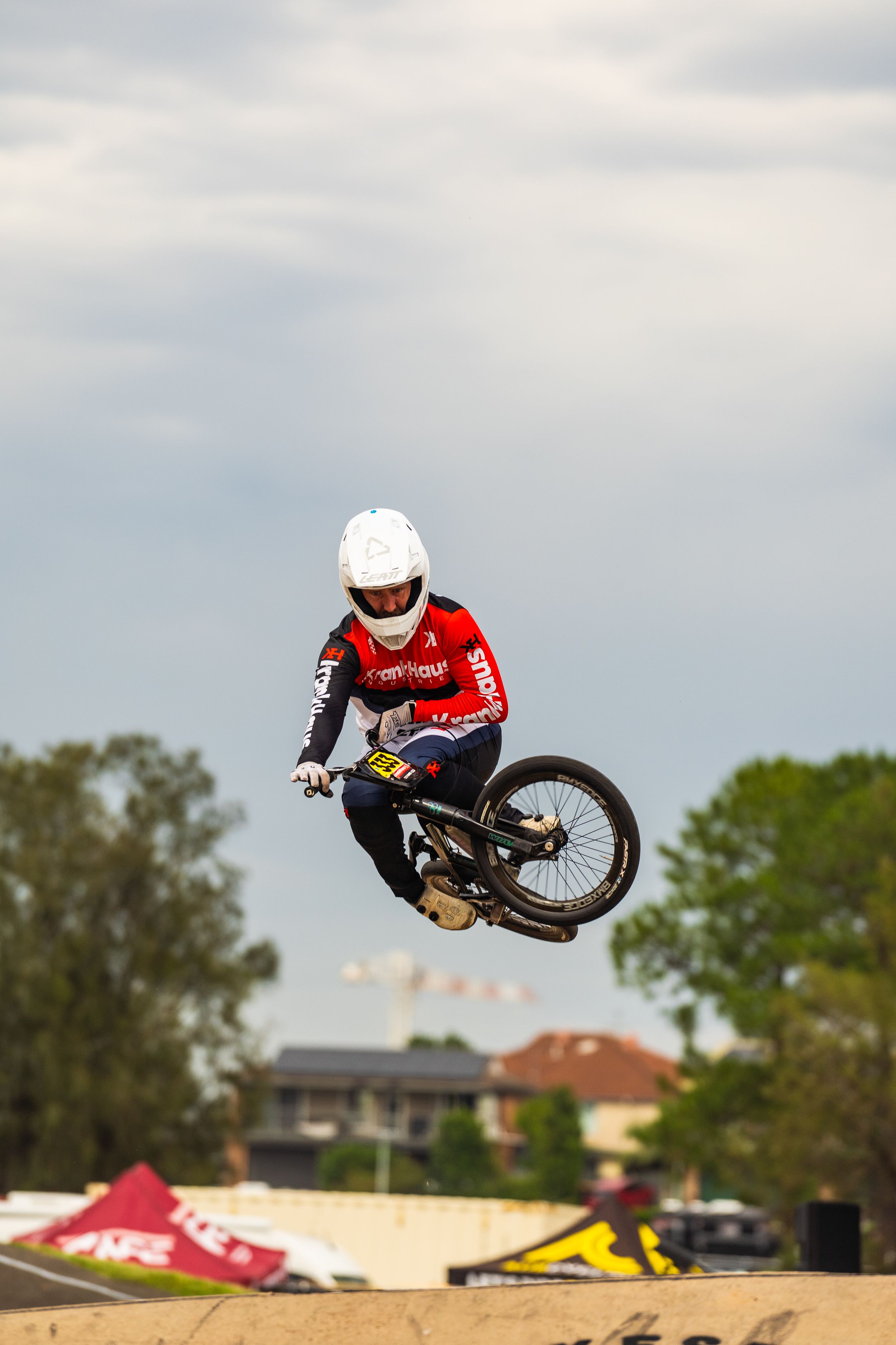 BMX rider performs aerial trick on dirt jump with residential buildings visible in background.