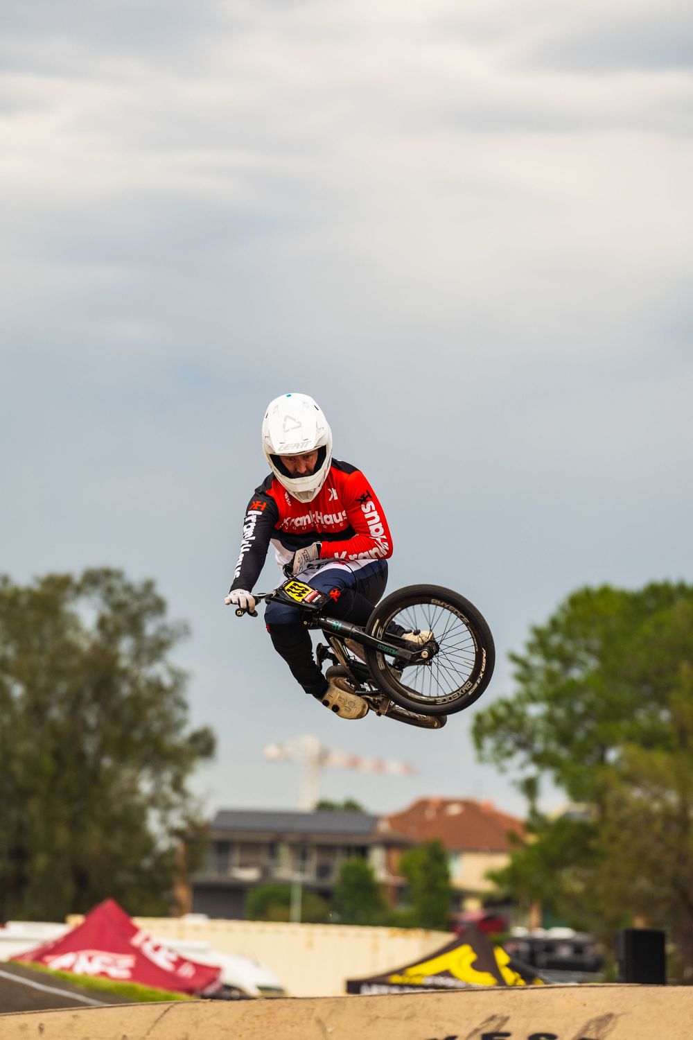 BMX rider performs aerial trick on dirt jump with residential buildings visible in background.