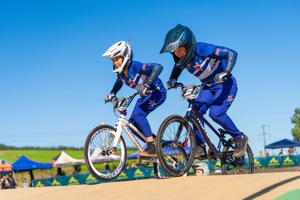 BMX riders compete on a dirt track, performing jumps and tricks against a bright blue sky.