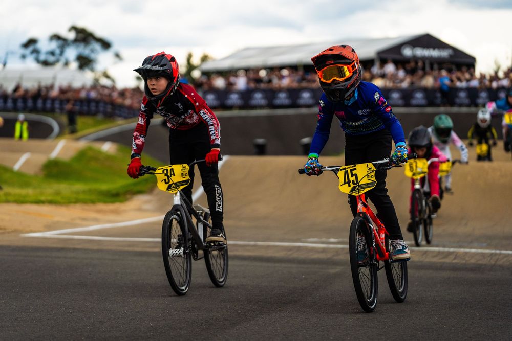 BMX riders performing tricks on a race track during a competitive event with spectators watching in the background.
