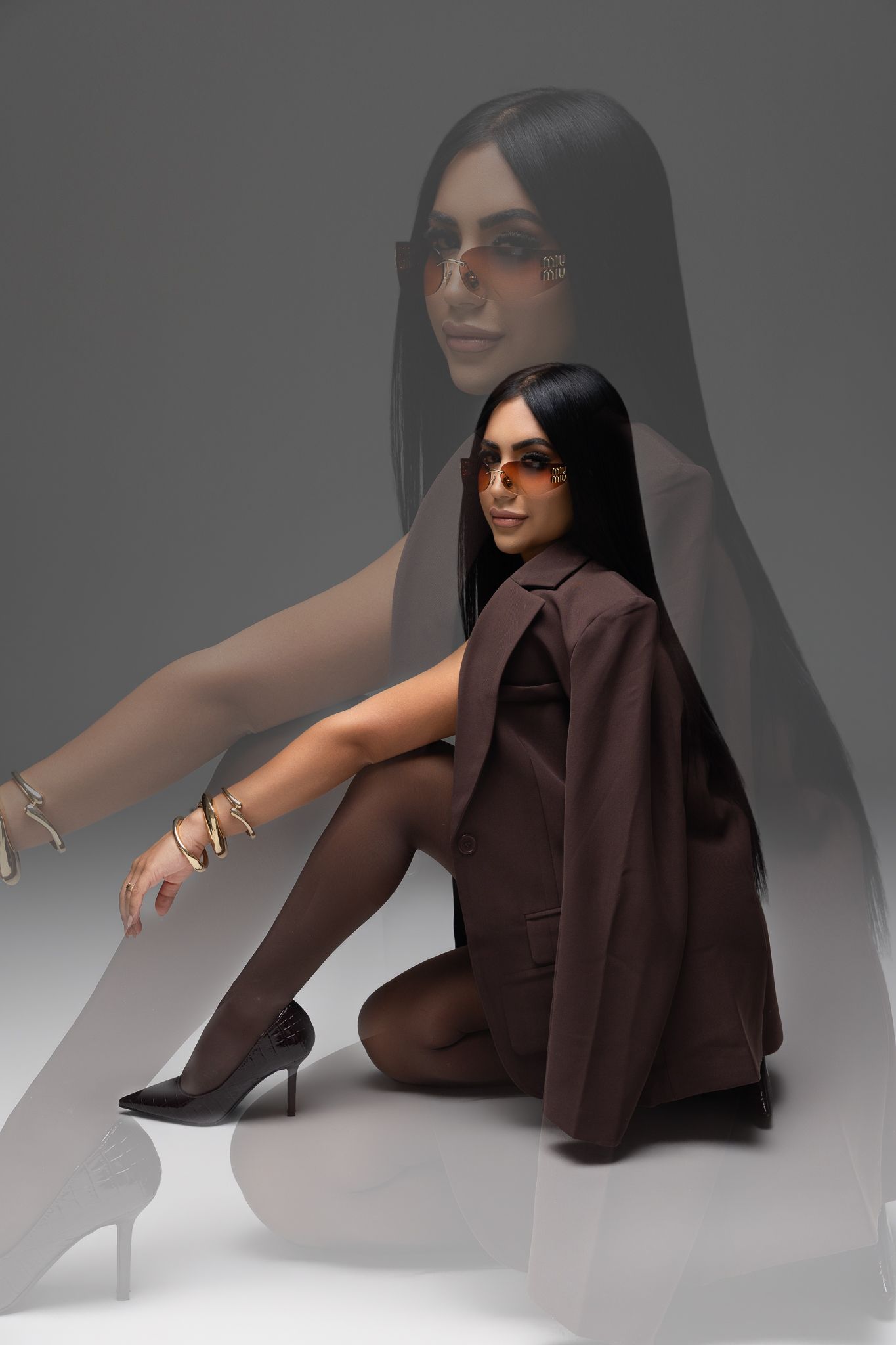 Stylish woman in brown blazer and orange sunglasses sitting on floor with reflection in background.