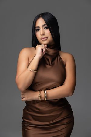 Woman in brown satin turtleneck dress with gold bracelets and ring poses elegantly against gray background.