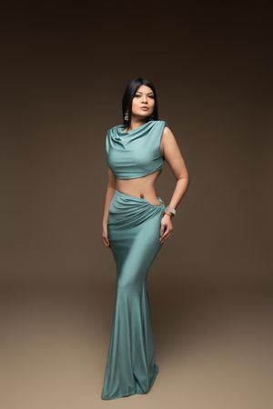 Woman in teal satin crop top and maxi skirt set posing elegantly against a warm brown studio background.