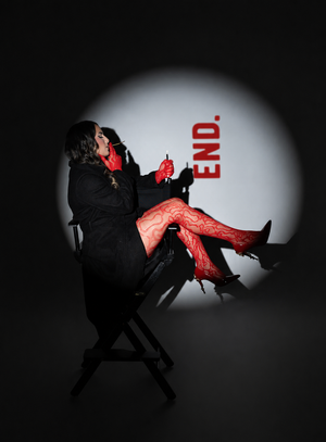 Woman in red lace tights and gloves sits in director's chair under spotlight with END text backdrop.