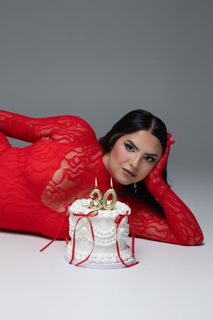 Woman in red lace outfit lying beside a white birthday cake with gold number 30 candles on gray background.