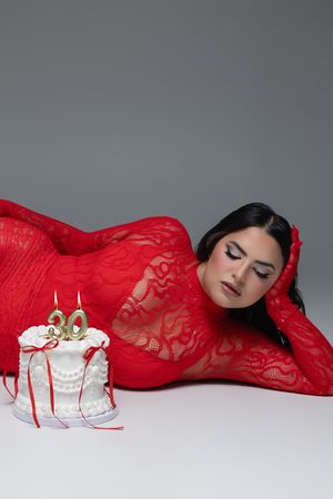 Woman in red lace bodysuit lying beside a white birthday cake with number 30 candles on gray background.
