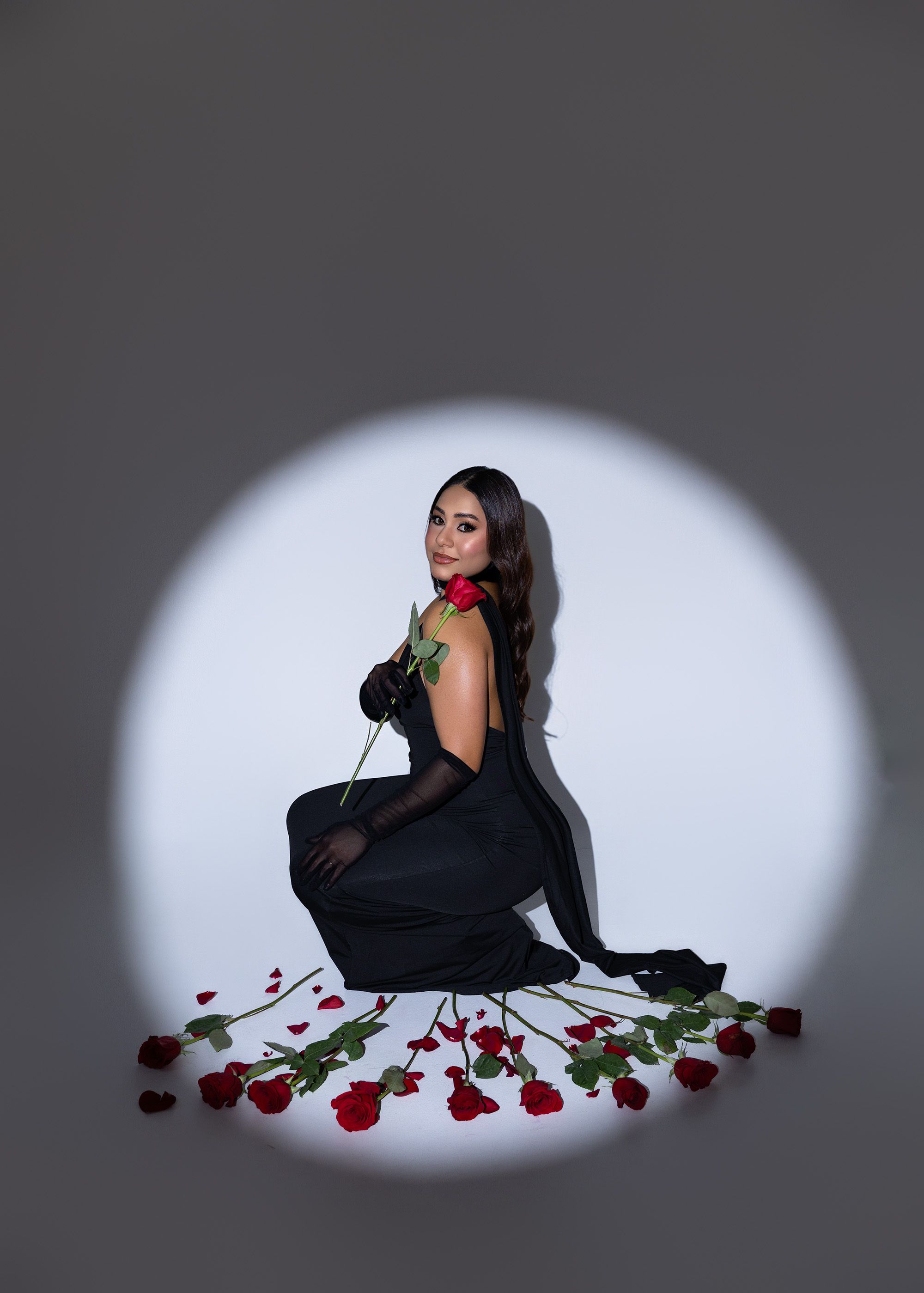 A dramatic spotlight illuminates a figure in black dress posed with scattered red roses on dark background.