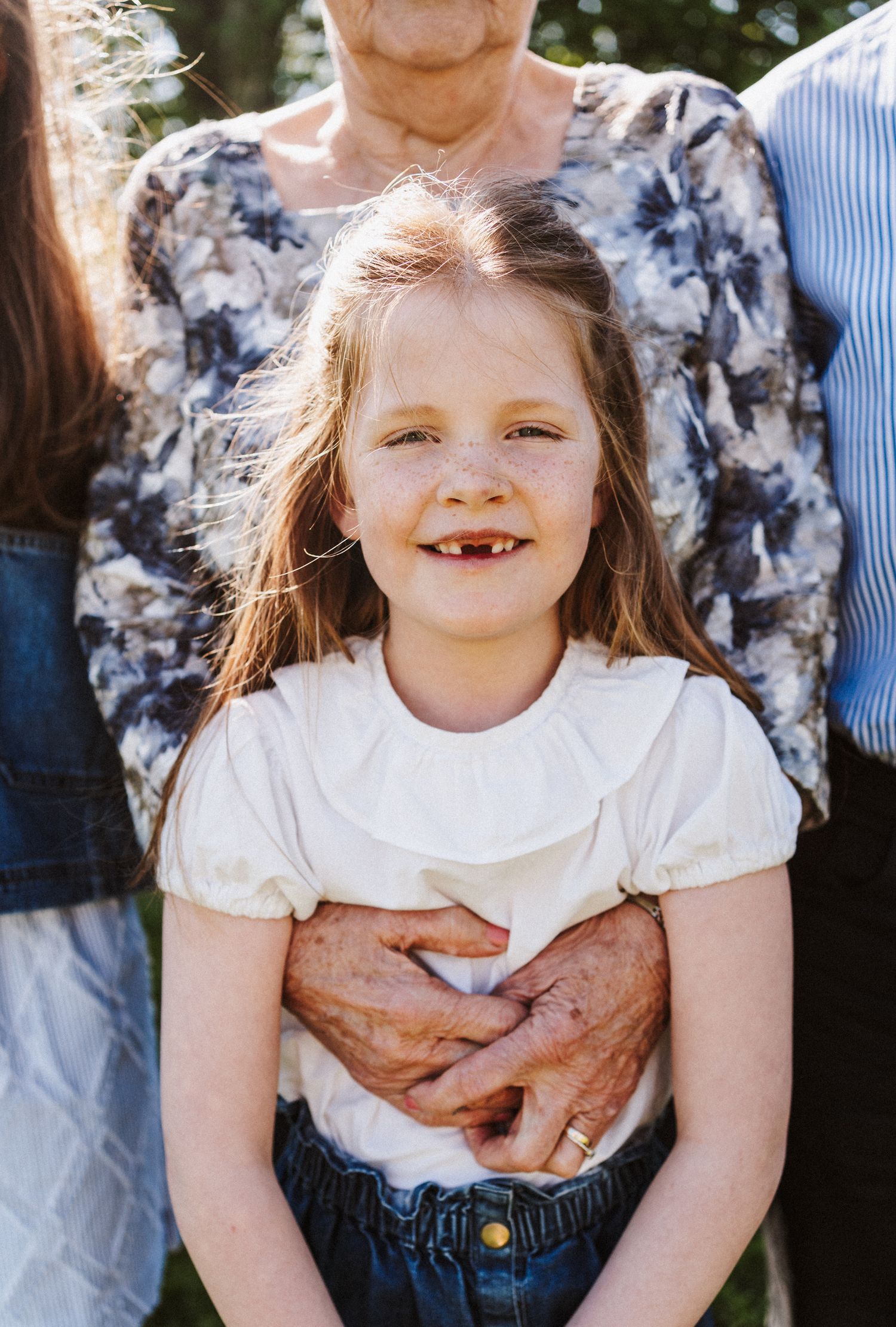 A young child wearing a white shirt and denim overalls smiles while being held in a playful moment outdoors.