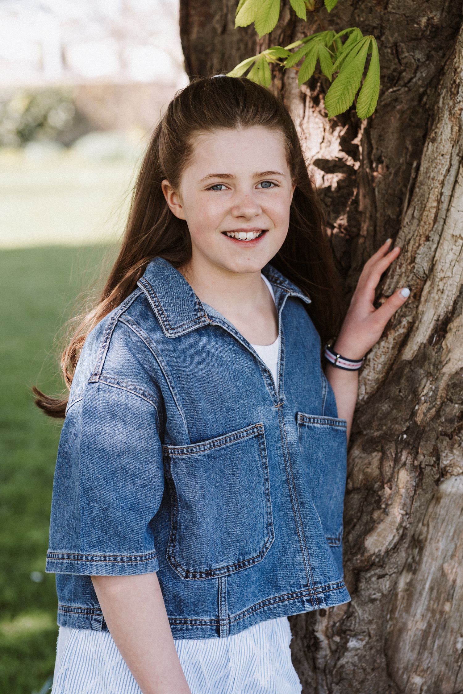 Person in denim jacket leaning against tree trunk with green leaves overhead.