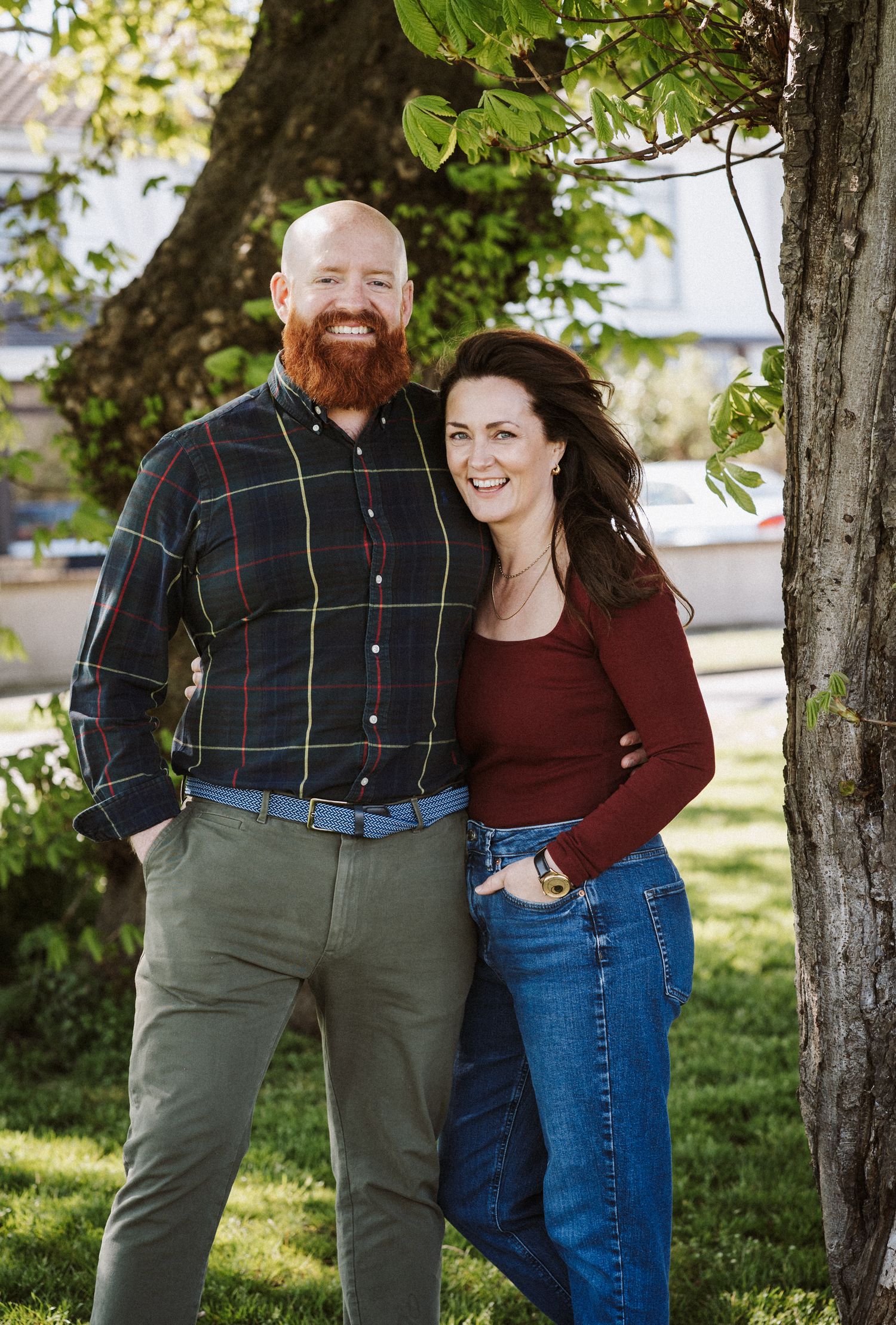 A couple stands together outdoors under a tree on a sunny day, wearing casual clothing and smiling at the camera.